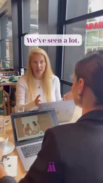 Two women talking at a café table by a window, laptops and coffee on the table